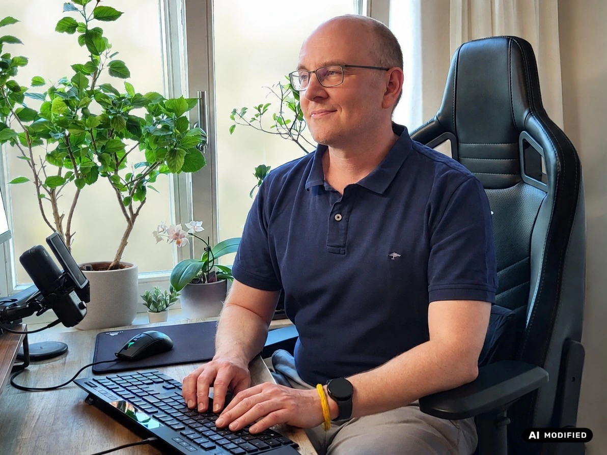 Raon Strohschein lächelnd bei der Arbeit an seinem Schreibtisch; Bildschirm und Fensterhintergrund KI-bearbeitet